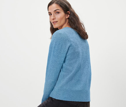 Frau sitzt auf einem Holzhocker und trägt eine hellblaue Strickjacke mit Wolle.