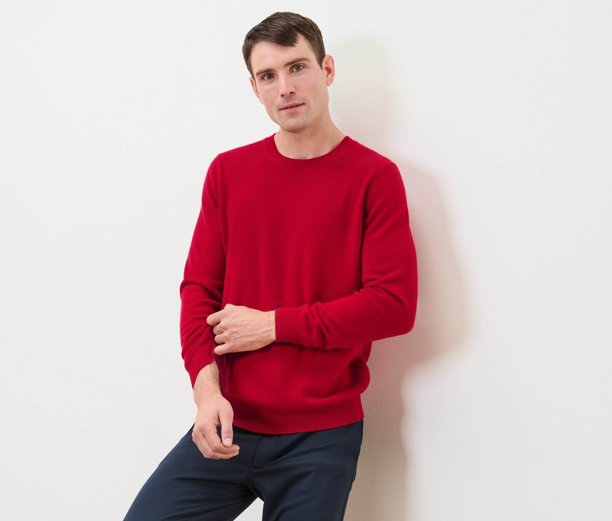 Mann lehnt an Wand, trägt roten Cashmere-Pullover mit Rundhalsausschnitt.