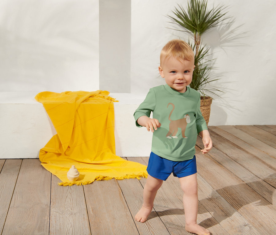 Kleiner Junge in einem Kinder-Shirt mit UV-Schutzfaktor 80 und blauen Badehosen steht auf einem Holzboden.