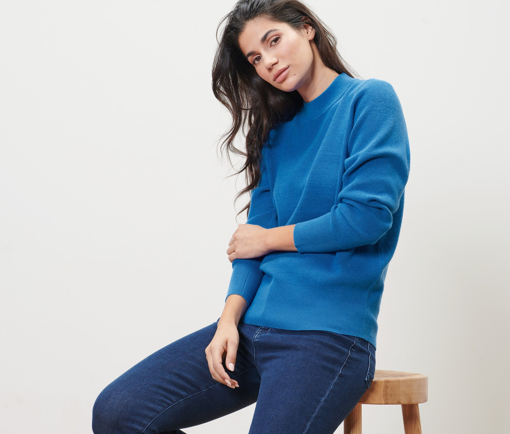 Frau sitzt auf einem Hocker und trägt einen blauen Feinstrickpullover und Jeans.