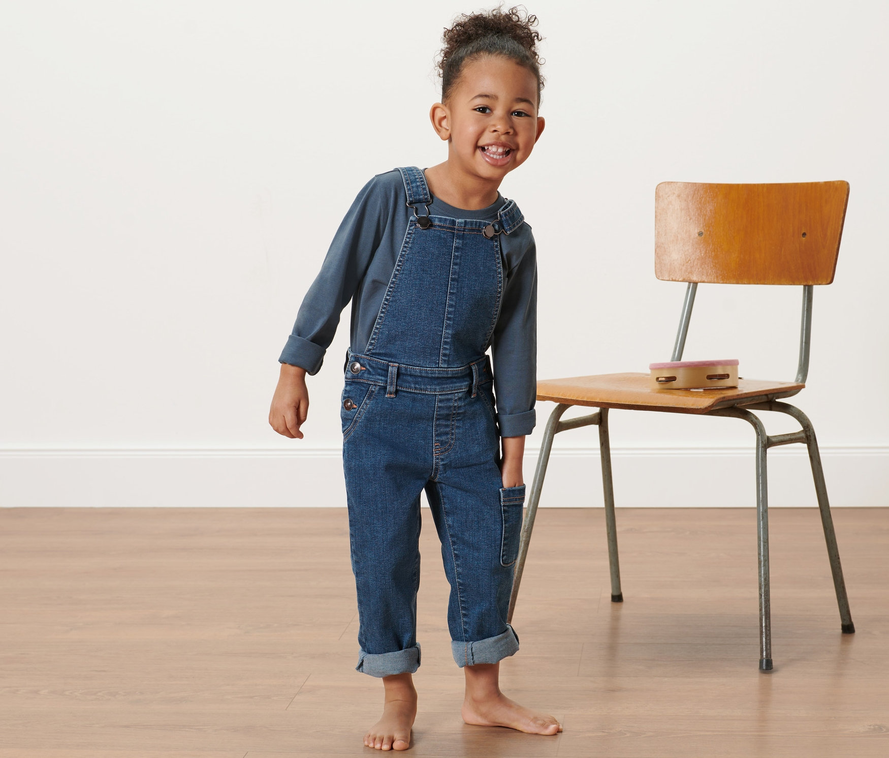 Lächelndes Mädchen, das eine Kleinkind-Denim-Latzhose trägt und neben einem Stuhl steht.