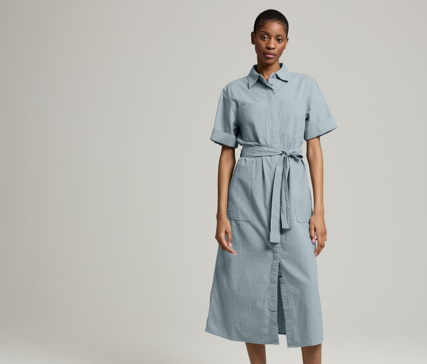 Eine Frau steht vor hellem Hintergrund und trägt ein NAH/STUDIO Seersucker-Kleid | Baumwolle, Stormy Grey und eine NAH/STUDIO Jeansjacke | ungefärbte Baumwolle und Turnschuhe.