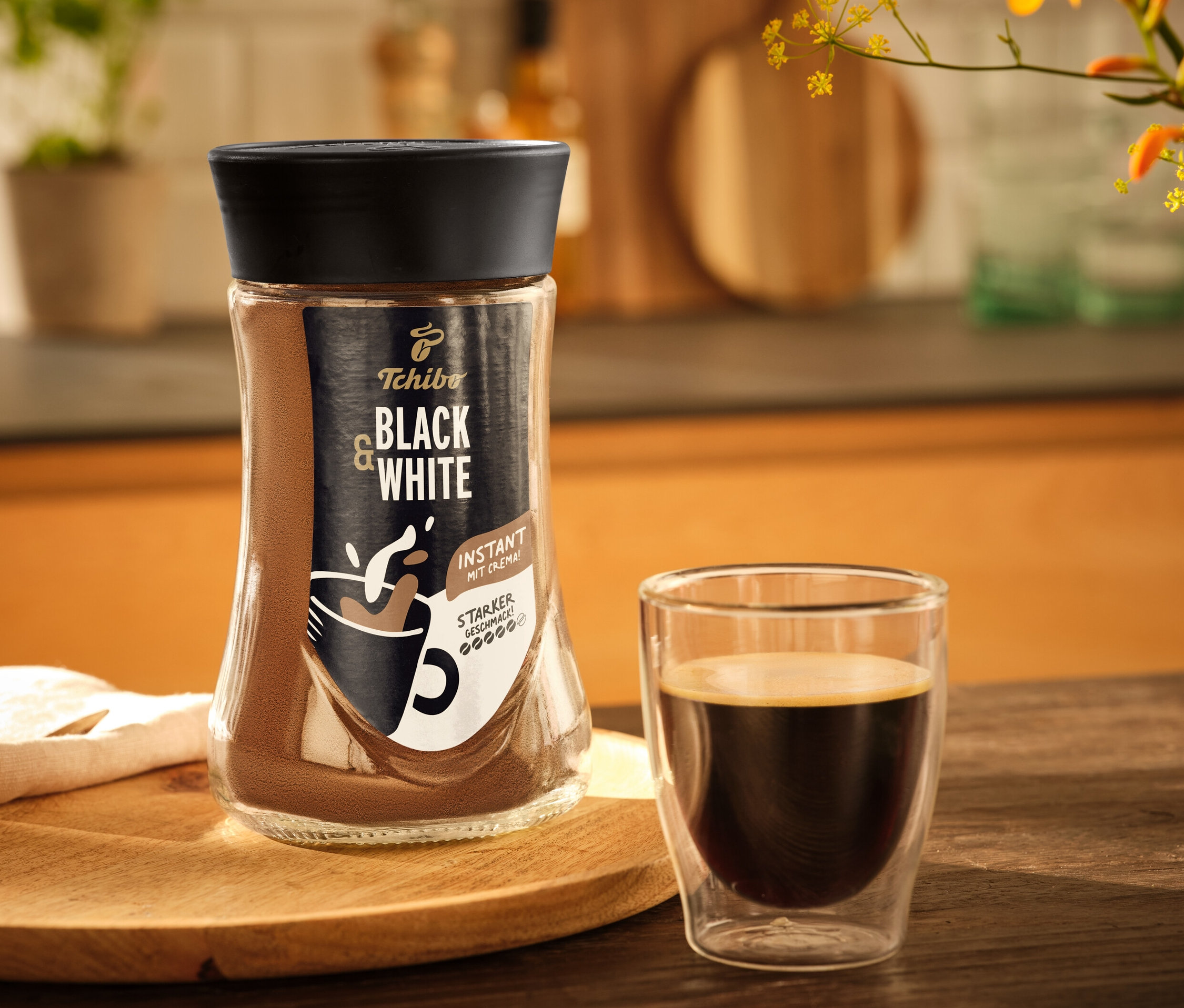 Auf einem Holztisch stehen ein Glas Tchibo Black & White Instantkaffee und eine Tasse Kaffee.