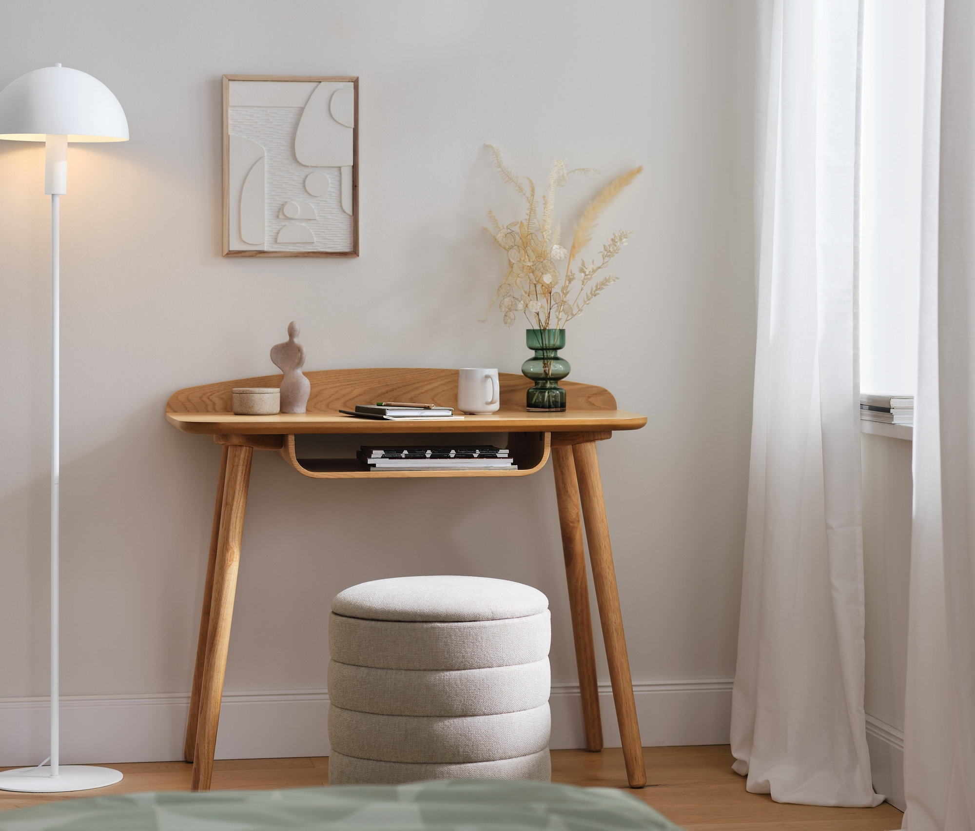 Eine weisse Stehleuchte aus Metall steht neben einem Schreibtisch. Unter dem Tisch steht ein 2-in-1-Pouf mit Stauraum.