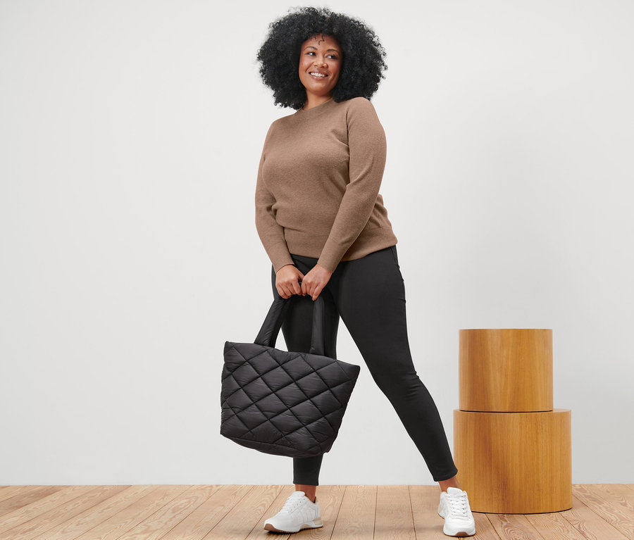 Frau posiert in braunem Strickpullover, schwarzen Leggings und weißen Turnschuhen und hält eine schwarze Stepptasche.