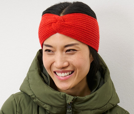 Eine Frau mit verschränkten Armen lächelt und trägt ein orangefarbenes Strick-Stirnband und eine grüne Daunenjacke.