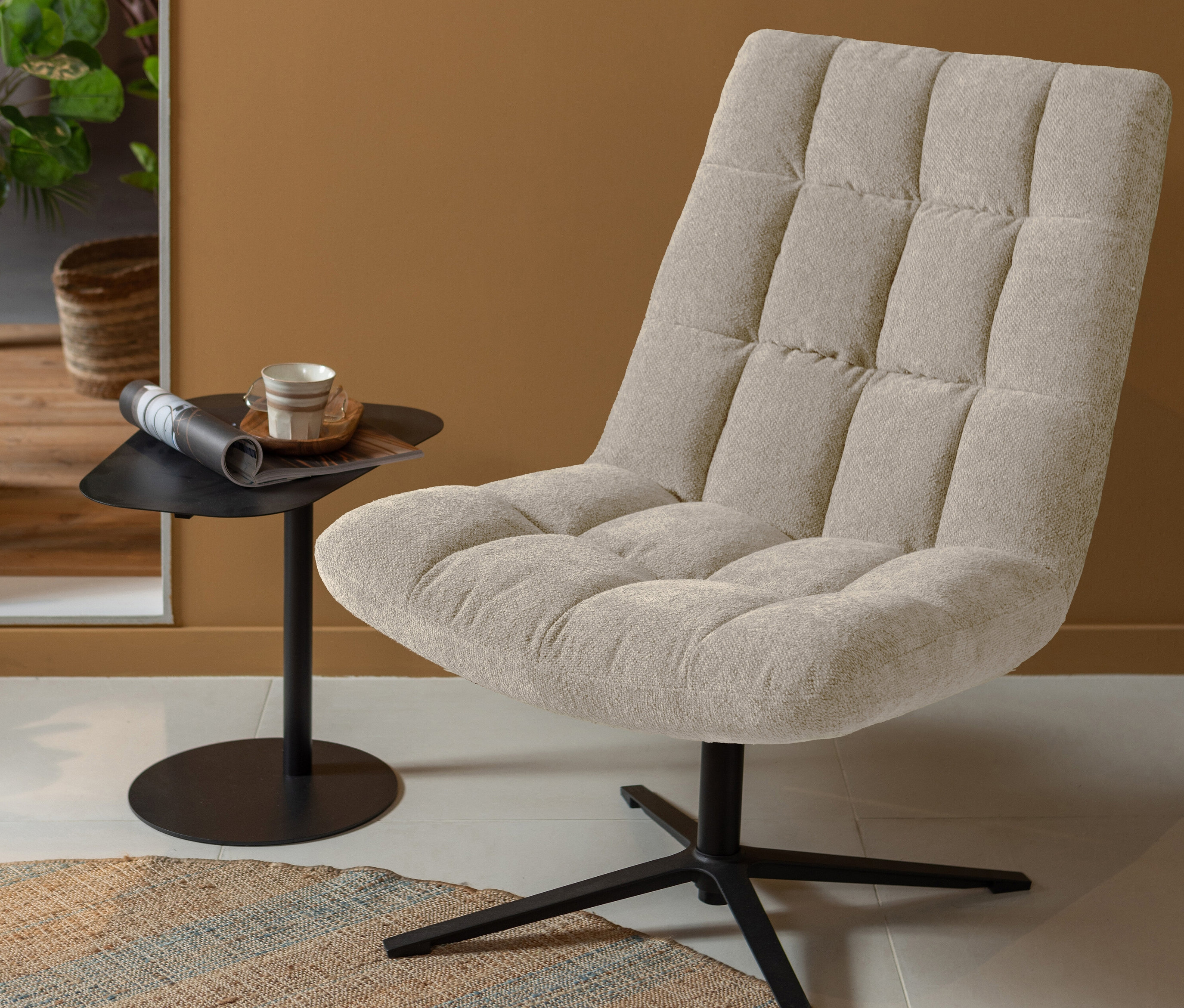 Ein sandfarbener Woood Drehsessel »Teddy« steht neben einem kleinen Tisch mit einer Tasse und einer Zeitung.