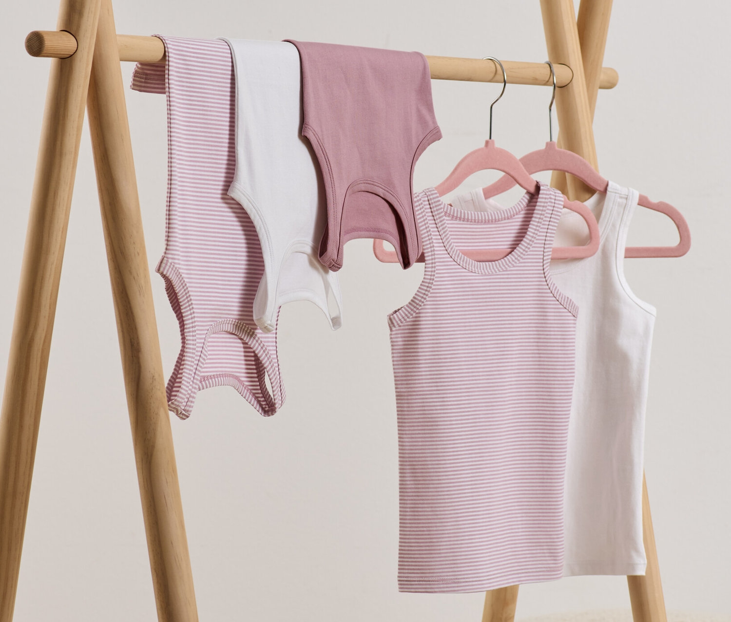 Auf einem hölzernen Kleiderständer hängen 5 Mädchen-Unterhemden, einige davon auf Kleiderbügeln.