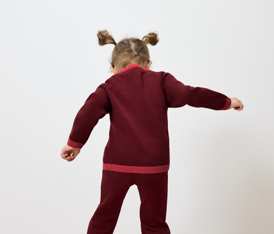 Ein Mädchen mit Zöpfen steht mit dem Rücken zur Kamera und winkt mit den Armen, es trägt einen burgunderroten Strickpullover und eine Hose.