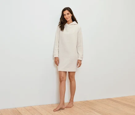 Eine Frau steht vor einer weißen Wand und trägt ein cremefarbenes Kapuzenkleid.
