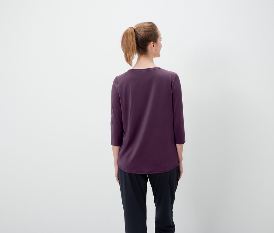 Rückansicht einer Frau mit braunen Haaren, die zu einem Pferdeschwanz zusammengebunden sind, in einem lila T-Shirt mit Dreiviertelärmeln und einer schwarzen Hose.