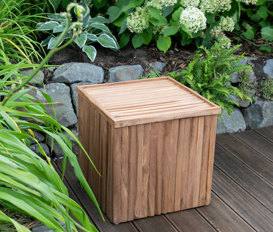 Ein brauner deVries Sitzhocker mit Stauraum steht auf einem Holzboden vor grünen Pflanzen.