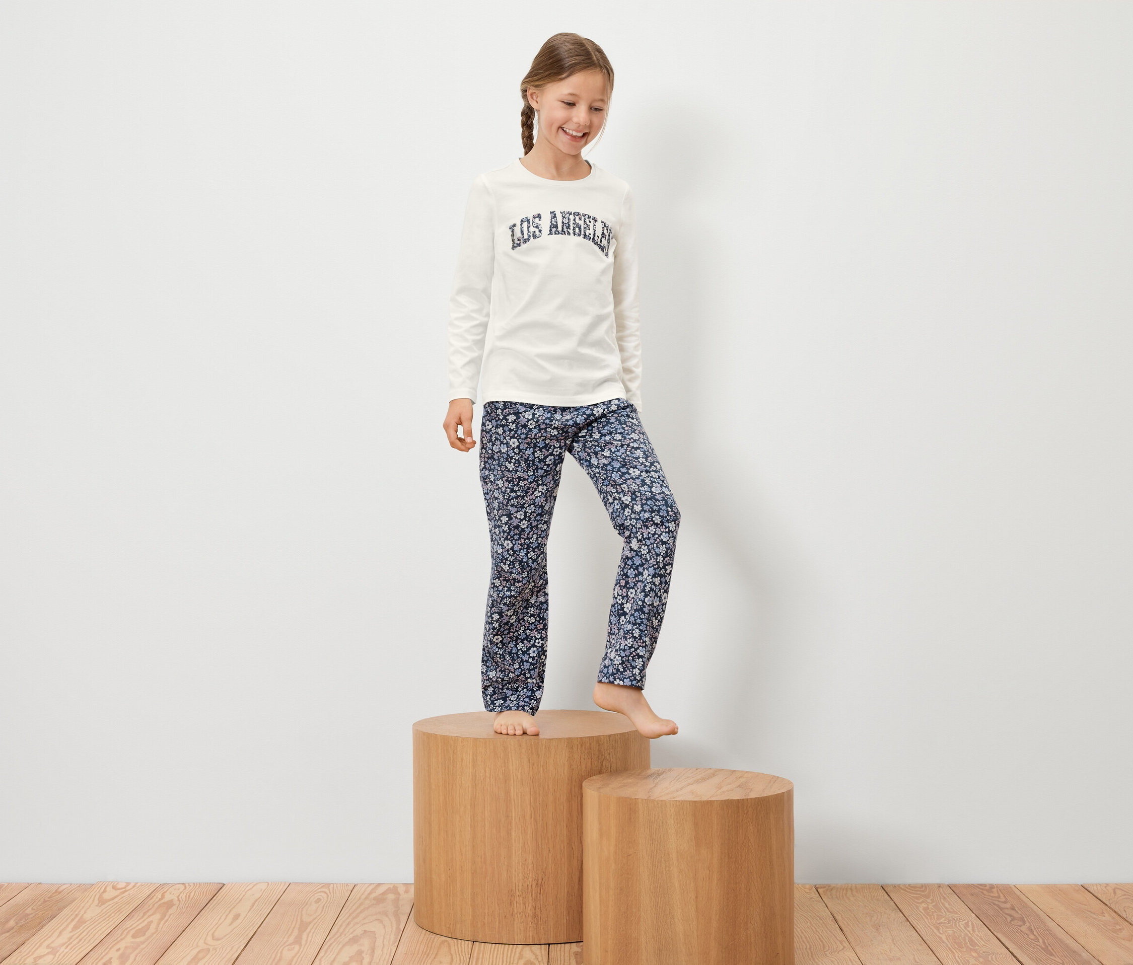 Mädchen mit Zopf steht auf einem Holzsockel in einem weißen T-Shirt mit Aufschrift und einer geblümten Hose.
