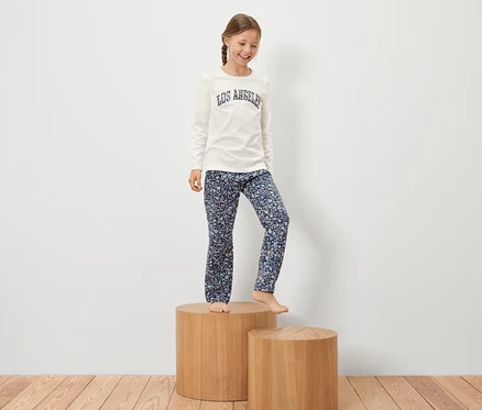 Mädchen mit Zopf steht auf einem Holzsockel in einem weißen T-Shirt mit Aufschrift und einer geblümten Hose.