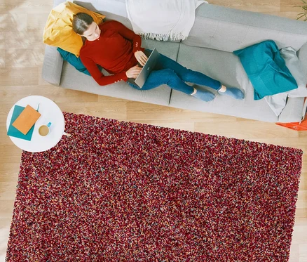 Eine Frau liegt auf einem grauen Sofa und benutzt einen Laptop. Im Vordergrund liegt ein rötlicher Teppich »Relic«, autumn spice.