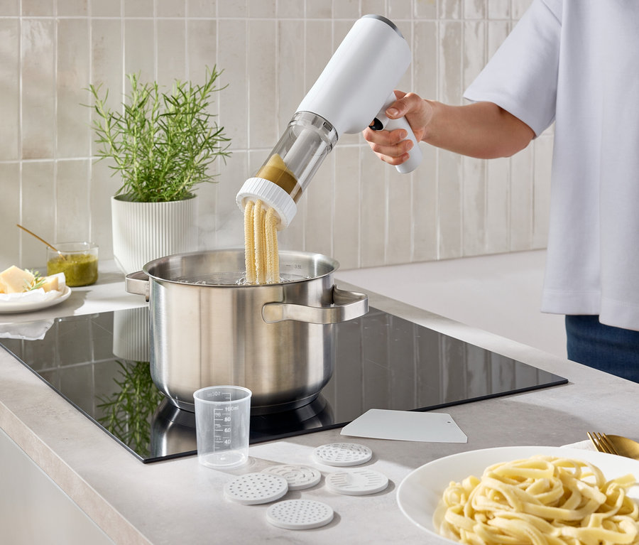 Eine Person gibt mit einer Nudelmaschine Nudeln in einen Topf mit Wasser. Auf der Arbeitsfläche steht ein Teller mit Nudeln.