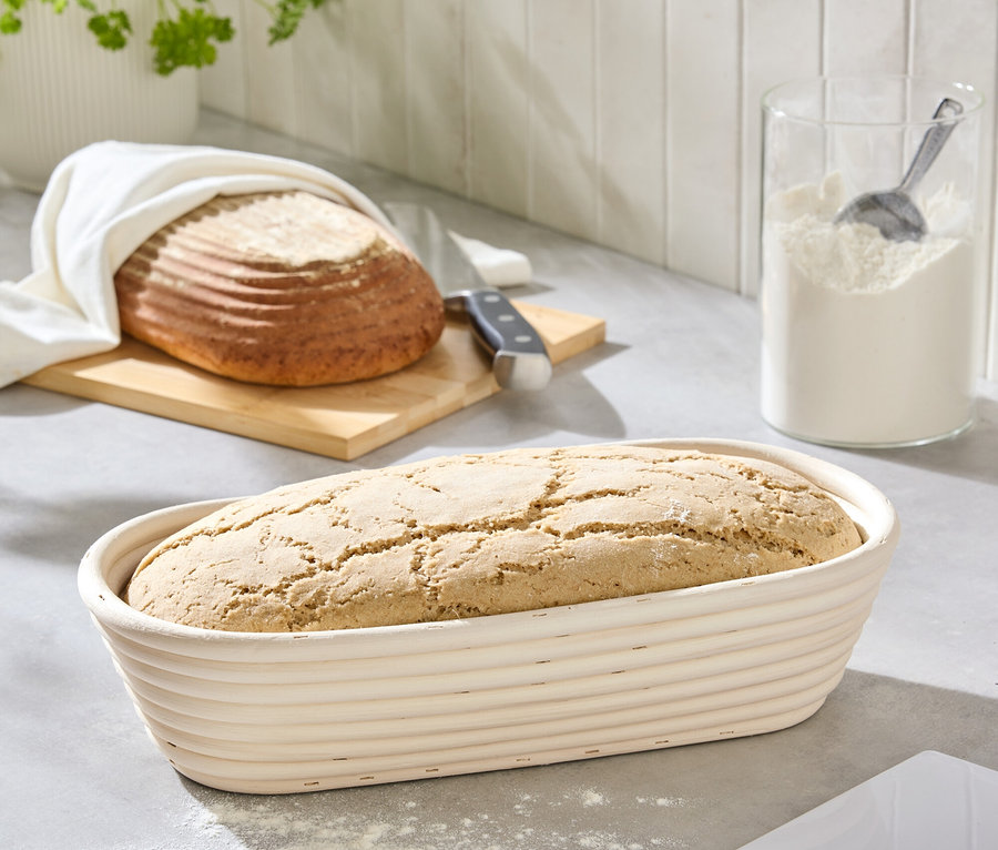 Brot im Gärkörbchen auf der Theke neben Mehl und Brot auf einem Schneidebrett.