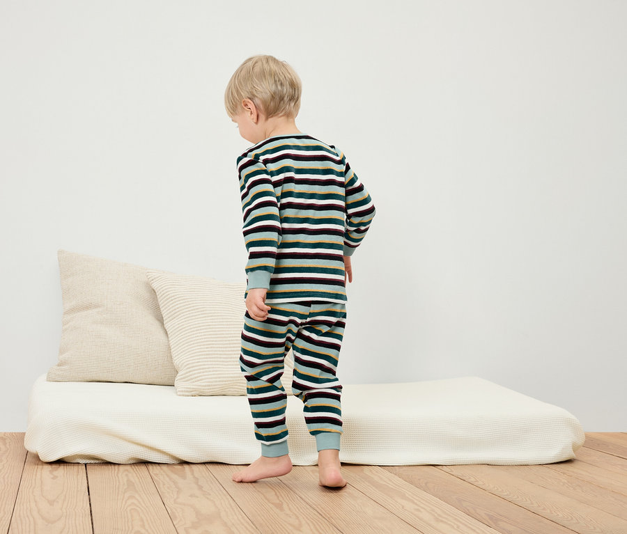 Junge steht in gestreiftem Pyjama auf dem Boden neben einem weißen Bett und Kissen.