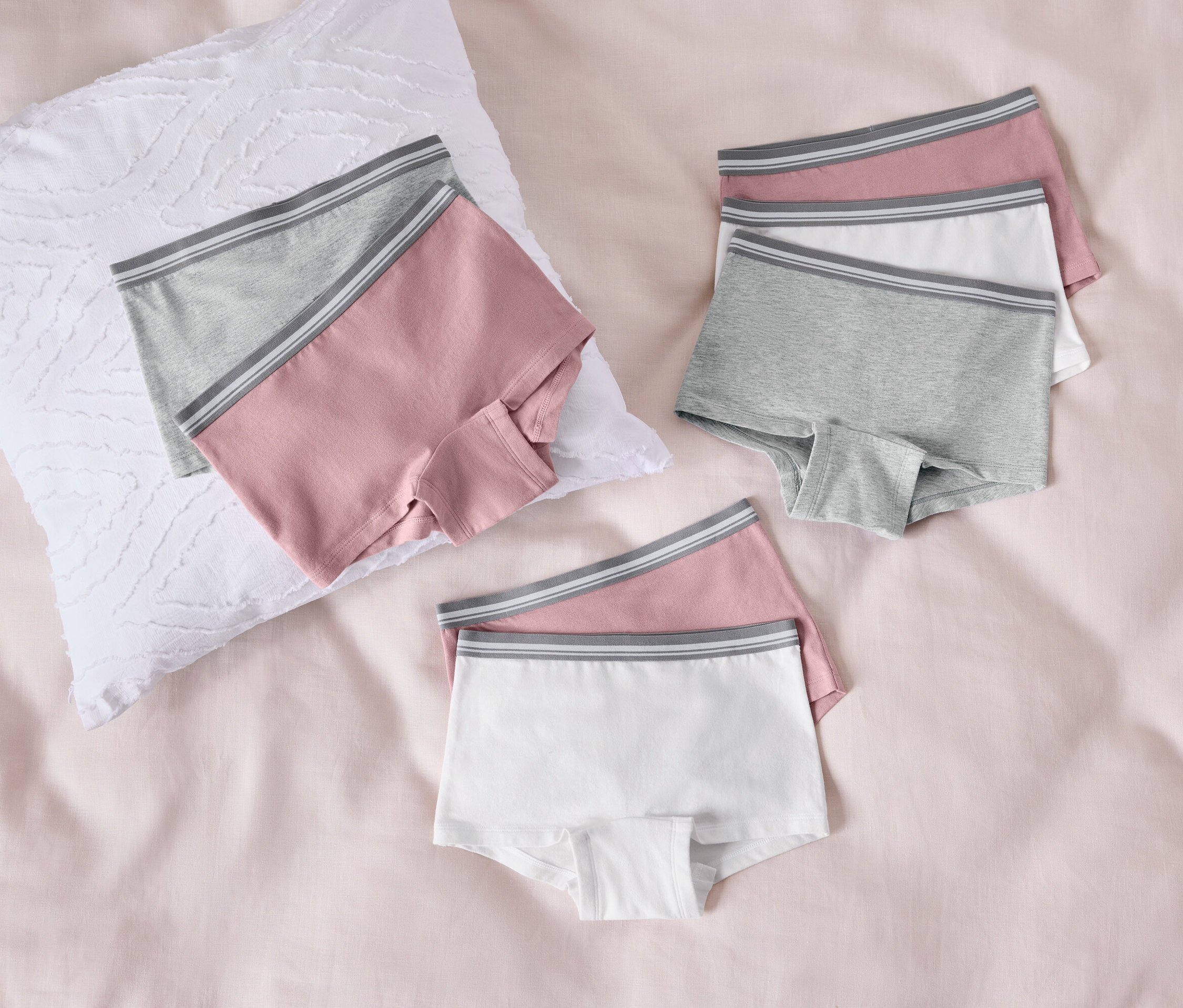 Drei Paar Damen-Boxershorts in Rosa, Grau und Weiß liegen auf einem rosa Bettlaken neben einem weißen Kissen.
