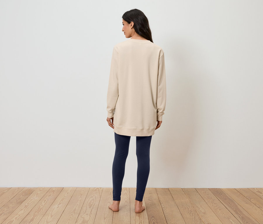 Frau steht mit dem Rücken zur Kamera und trägt ein beiges Sweatshirt und dunkelblaue Leggings.