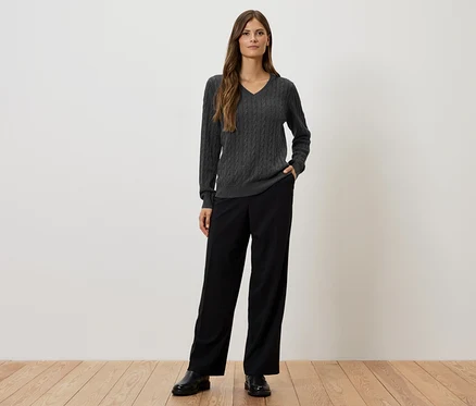 Eine Frau steht vor einer weißen Wand und trägt einen grauen Zopfmusterpullover, eine schwarze Hose und schwarze Schuhe.