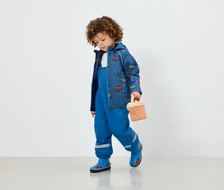 Ein Junge in blauer Latzhose, einer Jacke mit Autoaufdruck und blauen Gummistiefeln hält ein beige Spielzeug.