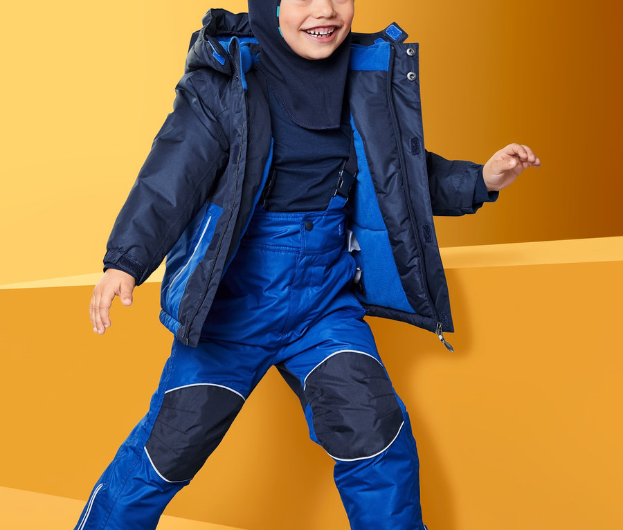 Ein Junge in einem blauen Schneekombi mit recyceltem Material posiert vor einem gelb-orangenen Hintergrund.