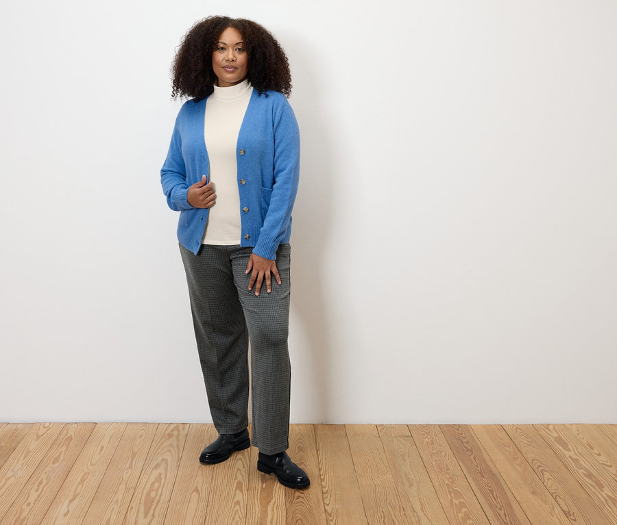 Frau steht auf Holzboden und trägt eine karierte Komfort-Jerseyhose, einen weißen Rollkragenpullover und eine blaue Strickjacke.