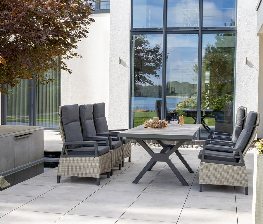 Gartenmöbel auf einer Terrasse. Ein Tisch und sechs Stühle mit grauen Kissen stehen auf Platten vor einem modernen Haus.