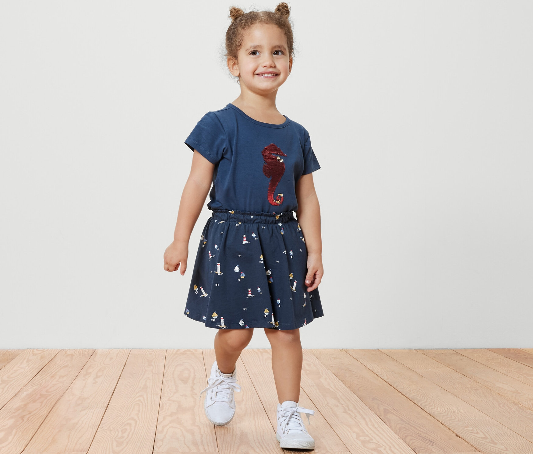 Lächelndes kleines Mädchen geht auf einem Holzboden und trägt einen Kinder-Jerseyrock mit Hose, maritim, und ein blaues T-Shirt mit einem Seepferdchen-Aufdruck.