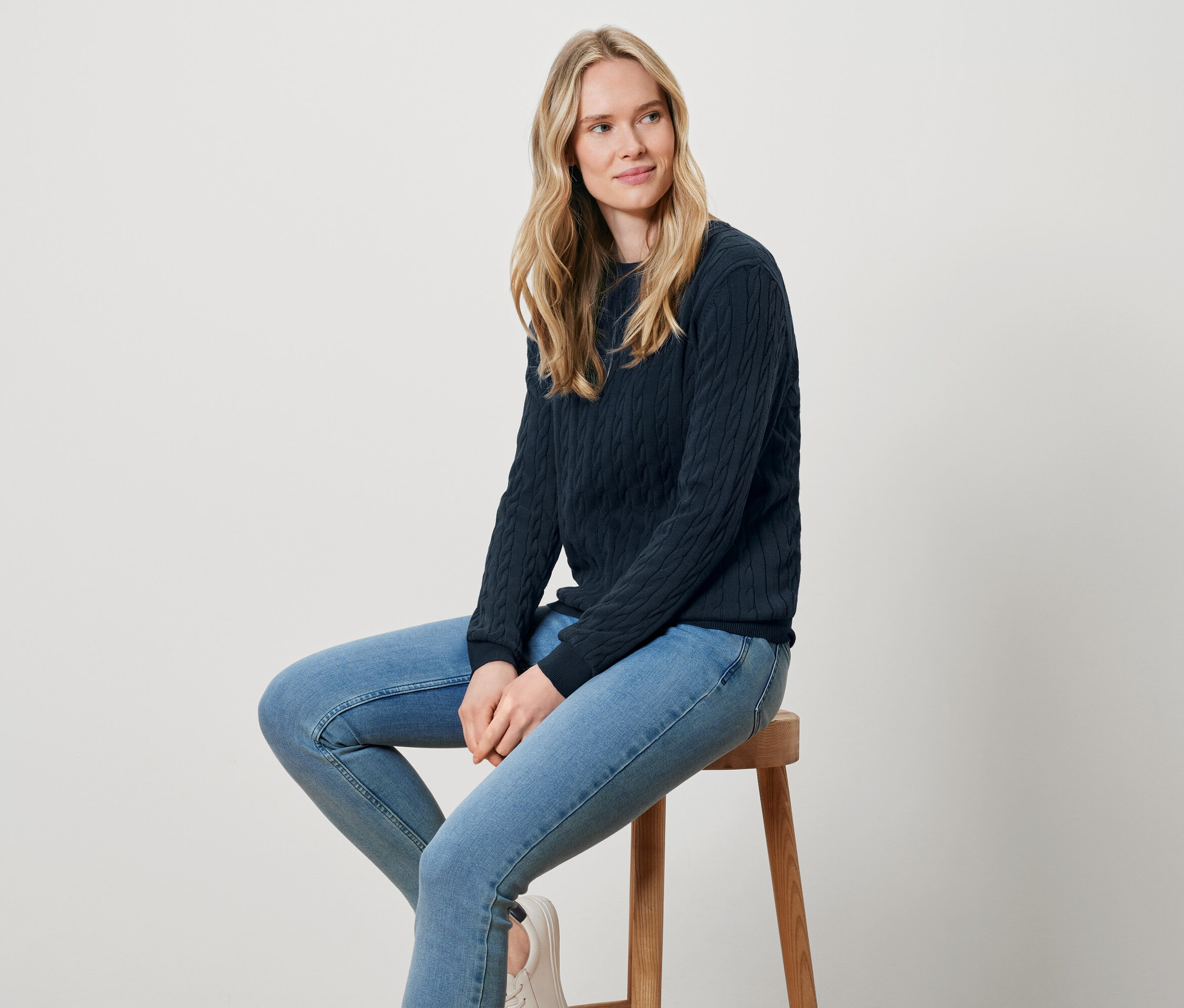 Eine blonde Frau sitzt auf einem Holzhocker und trägt einen dunkelblauen Strickpullover und Jeans.