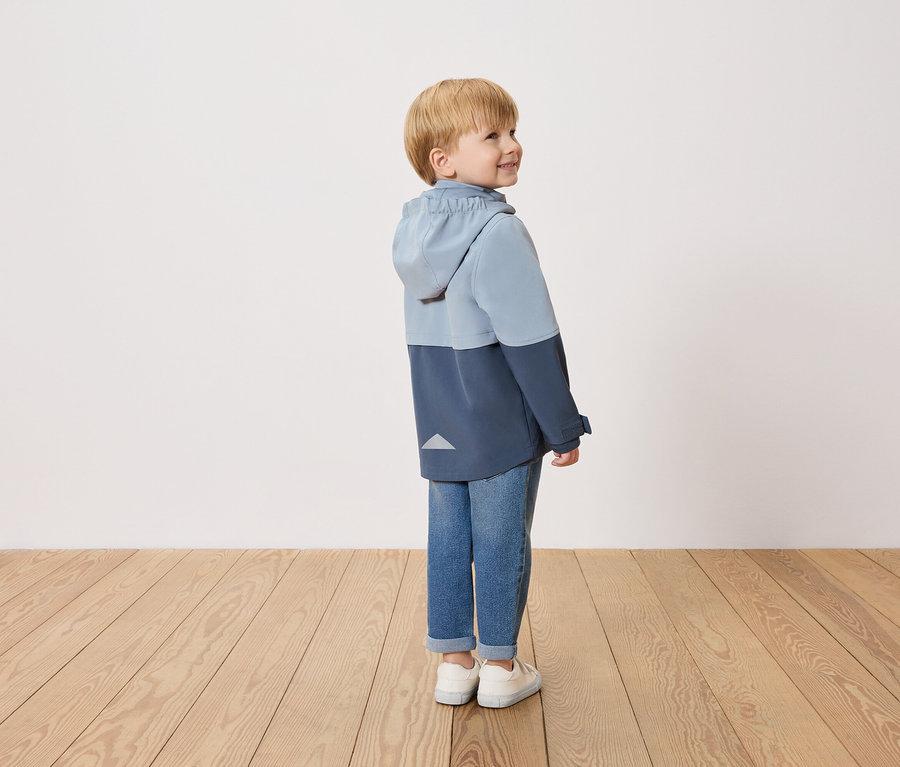 Ein lächelnder Junge steht seitlich und trägt eine zweifarbige blaue Jacke, Jeans und weiße Schuhe.