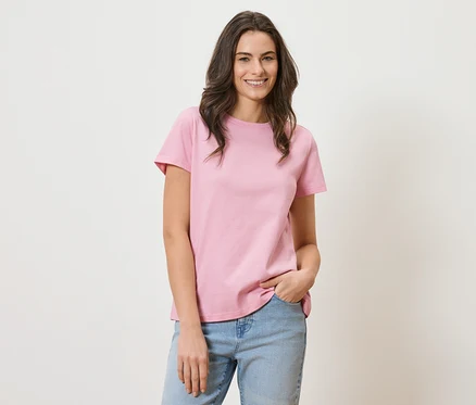 Frau mit langen braunen Haaren, die ein rosa T-Shirt und blaue Jeans trägt und lächelt.