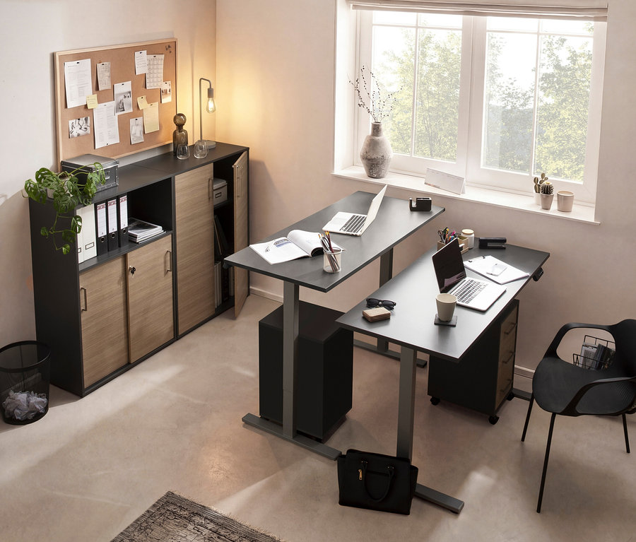 Büro mit zwei höhenverstellbaren Schreibtischen, Laptops, Büromaterial und einem Schrank mit Schiebetüren unter einer Korktafel.
