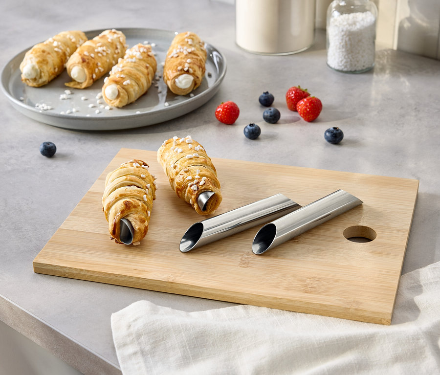 Auf einem Tisch liegen Windbeutel auf einem Teller und einem Schneidebrett, zusammen mit Beeren, Zucker und Metallformen.