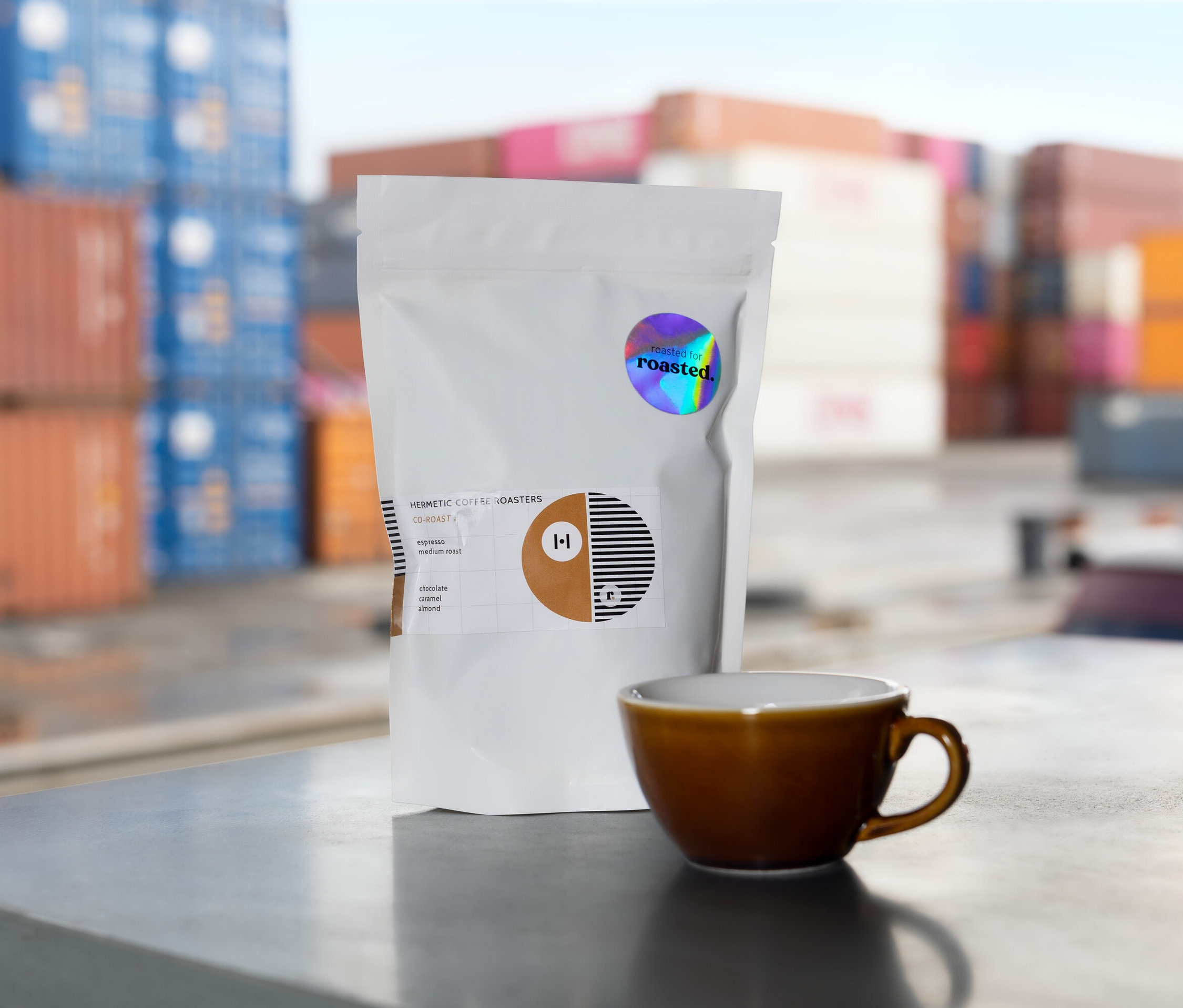 Eine weiße Packung Hermetic Coffee Roasters Kaffee steht neben einer braunen Tasse vor dem Hintergrund von Containern.