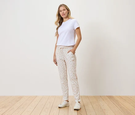 Frau steht vor einer Wand, trägt ein weißes T-Shirt, gemusterte Hose und weiße Turnschuhe.