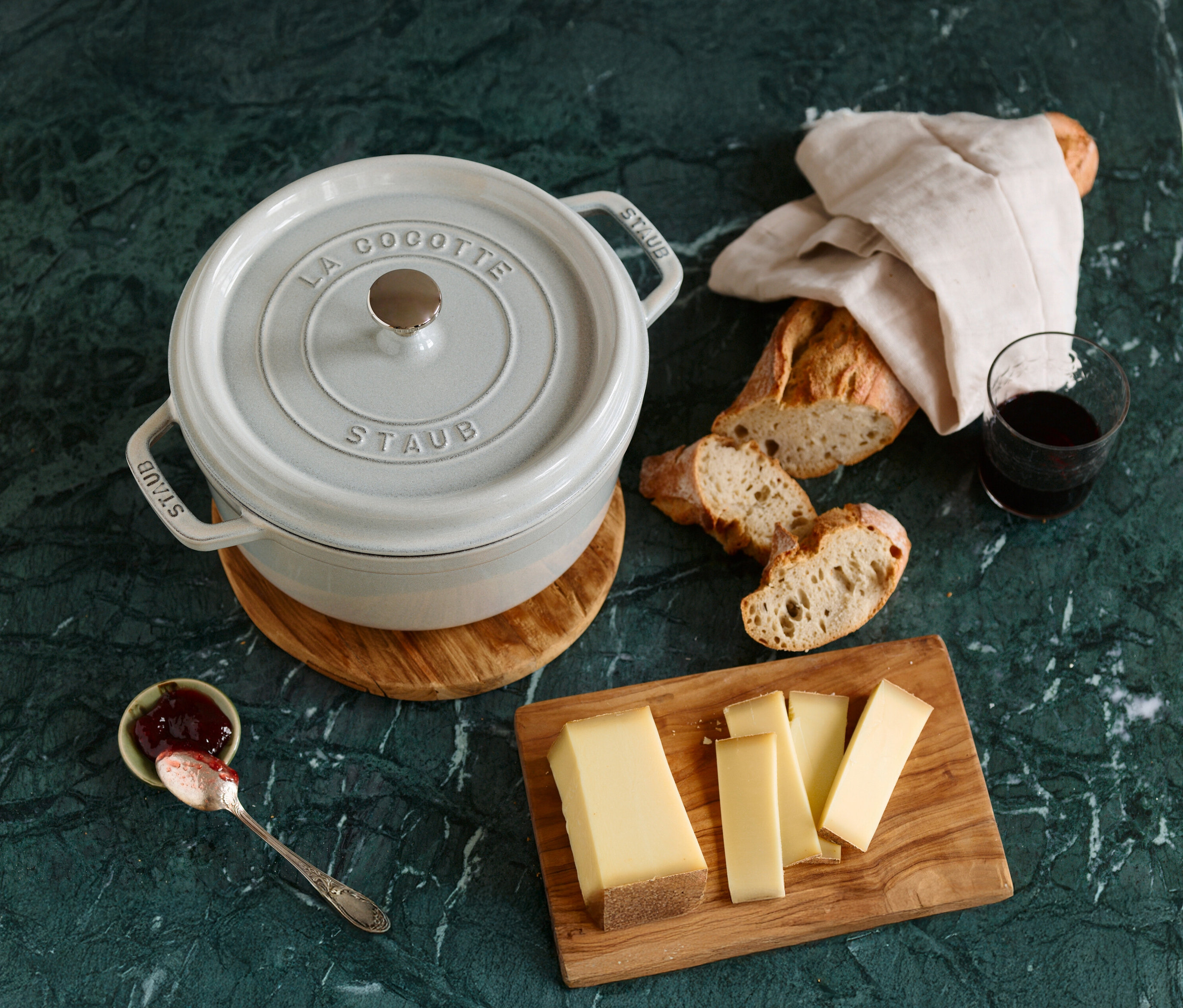 Ein gedeckter Tisch mit einem weißen STAUB »Cocotte 24« Kochtopf (ca. 3,7 l), Brot, Käse, Marmelade und einem Glas Wein auf einer grünen Marmorplatte.
