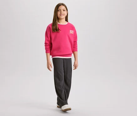 Mädchen mit pinkem Pullover mit Sonnenaufgangslogo und grauer Cordhose, dazu weiße Schuhe.