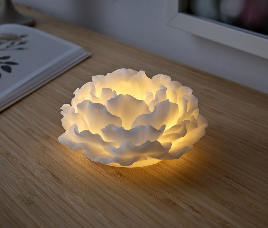 Eine weiße, blütenförmige Lampe leuchtet auf einem Holztisch. Daneben liegt ein aufgeschlagenes Buch.