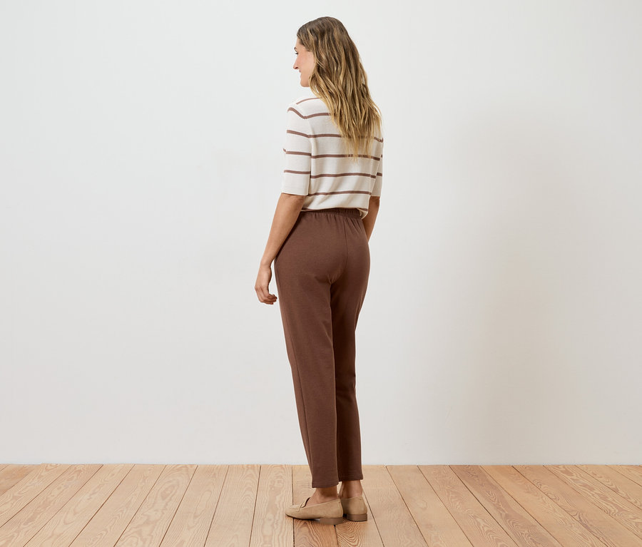 Frau von hinten in brauner Hose, gestreiftem Oberteil und beigen Schuhen vor einer weißen Wand.