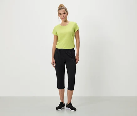 Frau steht mit grünem T-Shirt, schwarzen Dreiviertelhosen und schwarzen Turnschuhen vor einem weißen Hintergrund.