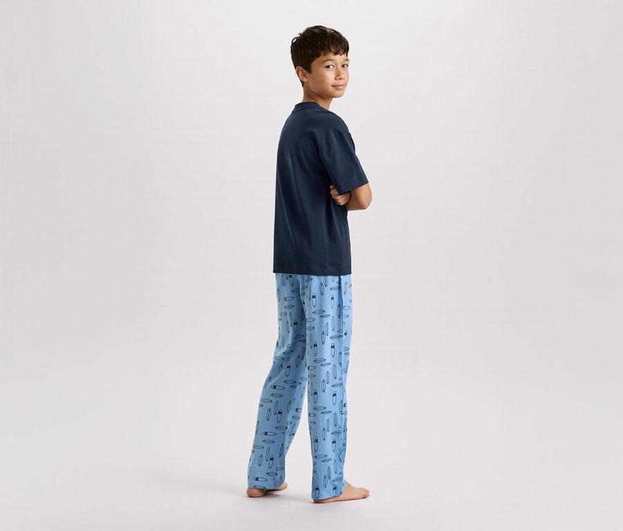 Ein Junge steht mit verschränkten Armen da, er trägt ein marineblaues T-Shirt und eine hellblaue Pyjamahose mit Surfbrettmuster.