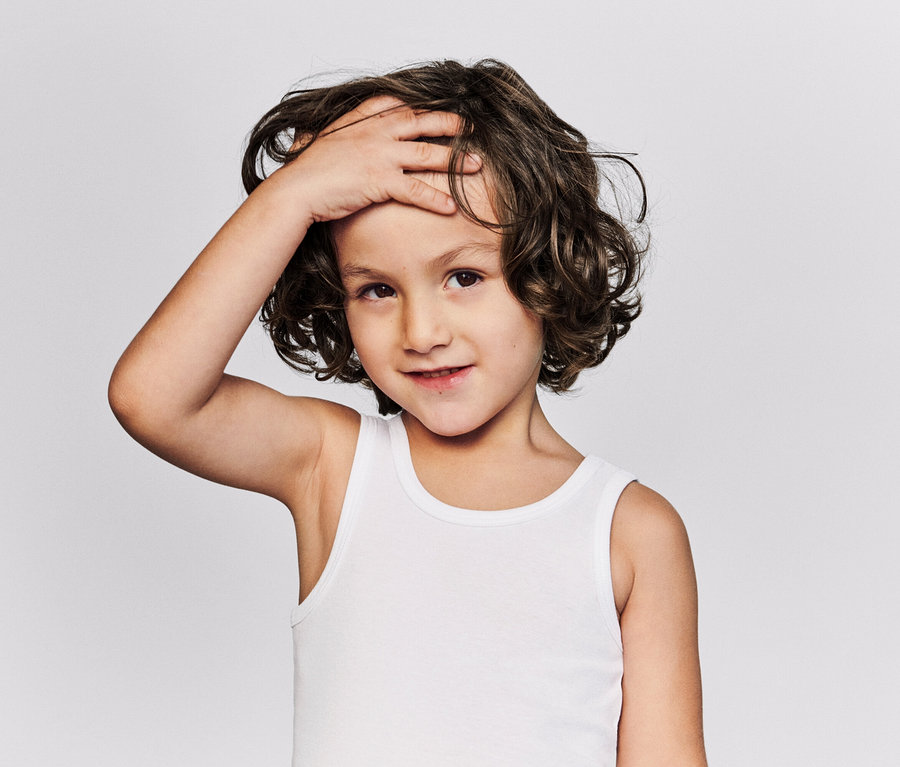 Porträt eines Kindes mit lockigem braunem Haar und einem weißen Unterhemd, das eine Hand auf die Stirn legt.