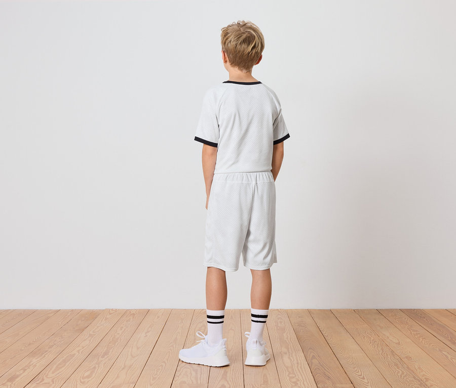 Ein Junge steht mit dem Rücken zur Kamera, er trägt ein weißes T-Shirt mit schwarzen Borten und passende Shorts, dazu weiße Socken mit schwarzen Streifen und weiße Turnschuhe.