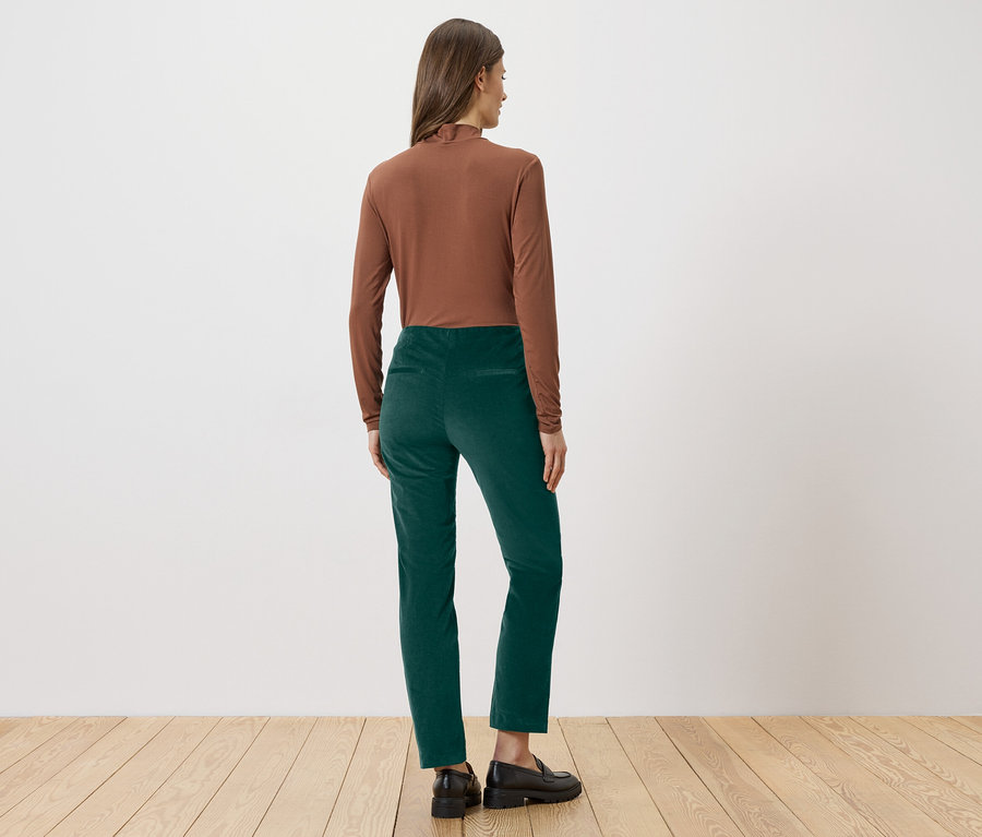 Frau von hinten in braunem Rollkragenpullover, grüner Hose und schwarzen Schuhen vor weißer Wand und Holzboden.