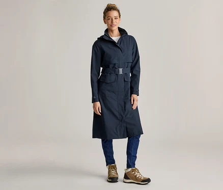 Frau mit Dutt trägt einen langen, marineblauen Regenmantel mit Gürtel, blaue Jeans und braune Wanderschuhe vor einem hellgrauen Hintergrund.