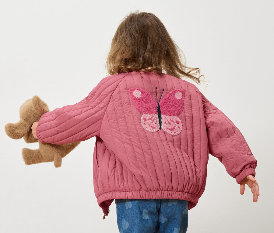 Rückansicht eines Kindes mit Teddybär, das eine rosa gesteppte Jacke mit Schmetterlingsapplikation trägt.