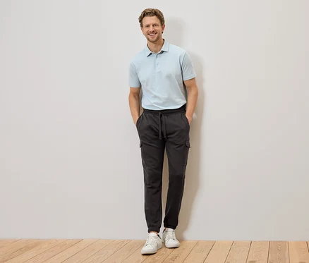 Ein Mann mit hellblauem Poloshirt und grauer Cargohose steht mit den Händen in den Hosentaschen vor einer Wand.