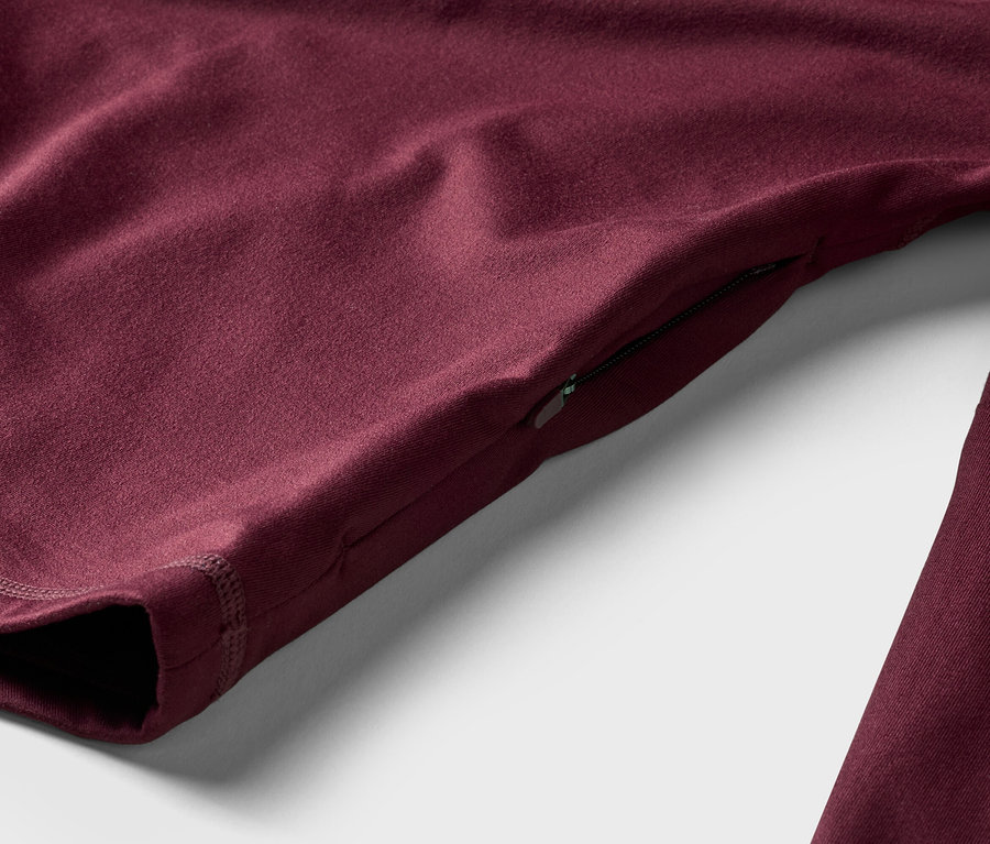 Nahaufnahme eines burgunderroten Langarmshirts mit Reißverschlusstasche am Ärmel, liegend auf einem weißen Hintergrund.